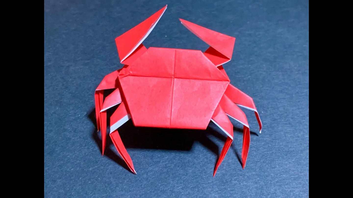 赤い折り紙で作ったカニが紺色の背景の上に置かれている。2本のハサミと左右4本ずつの足が見える
