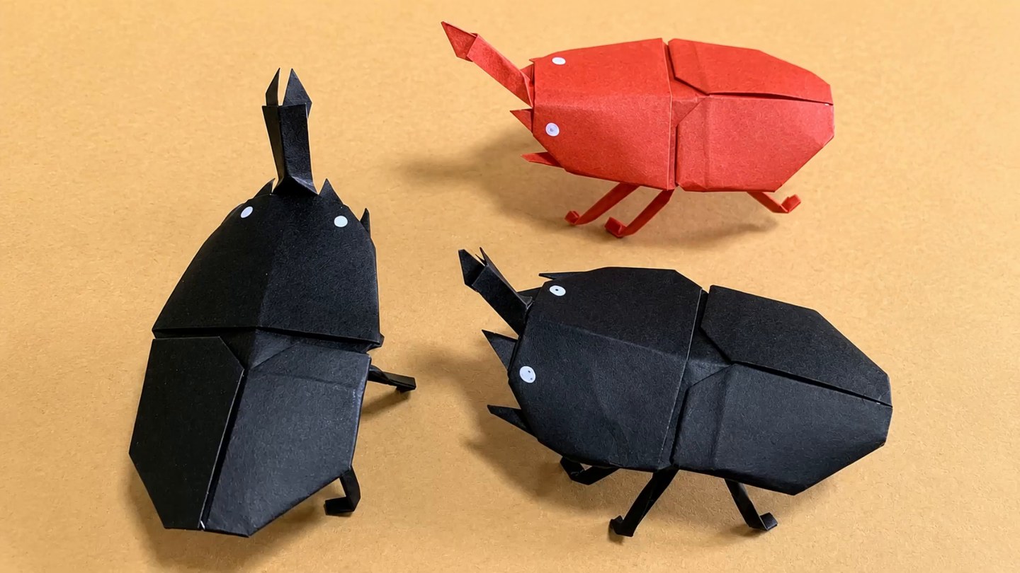 黒い折り紙で折ったカブトムシ2匹と赤い1匹がオレンジ色の背景に並び、角と白い点の目、折り曲げた足が見える