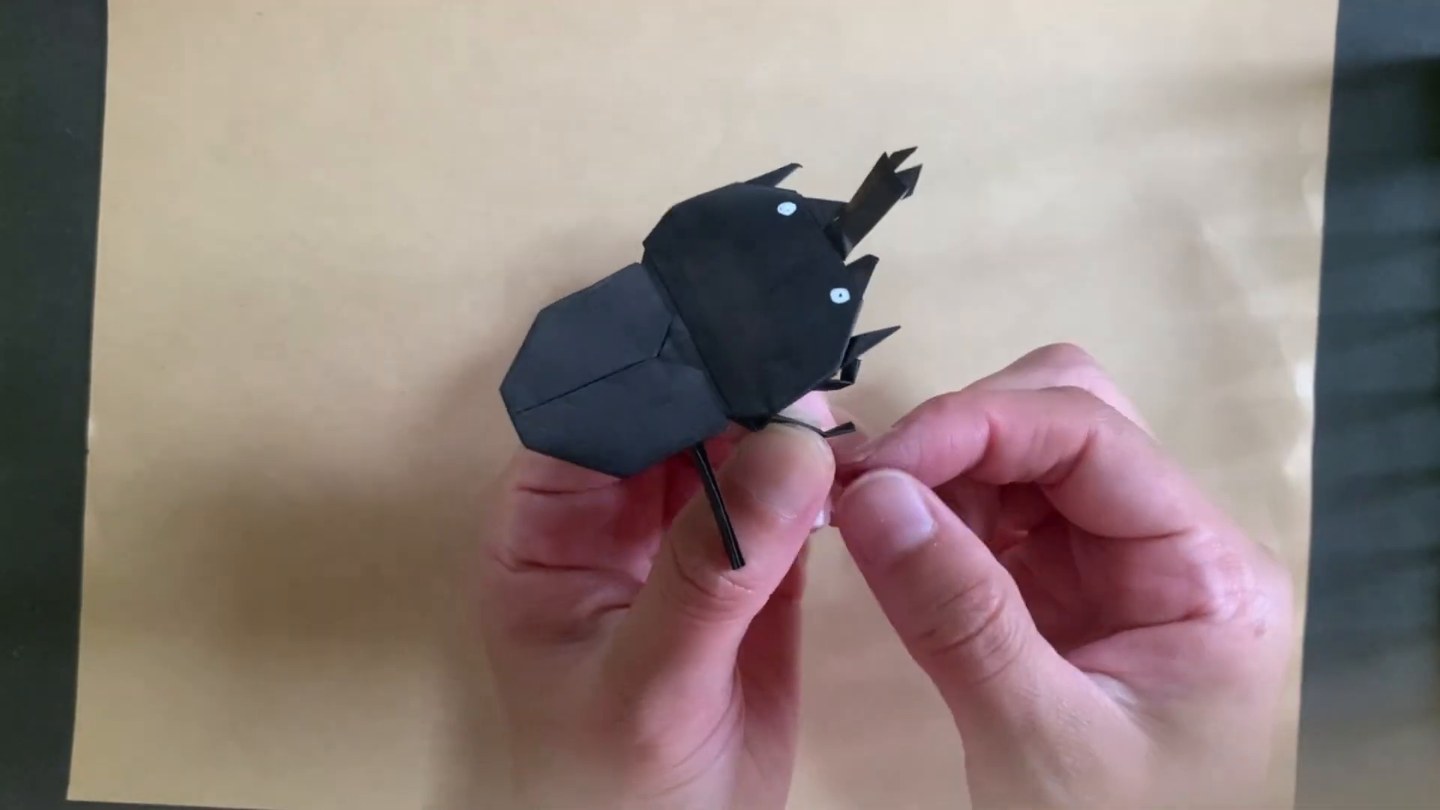 角と白い点の目が付いた黒いカブトムシの折り紙を両手で持ち、足を付けているところ