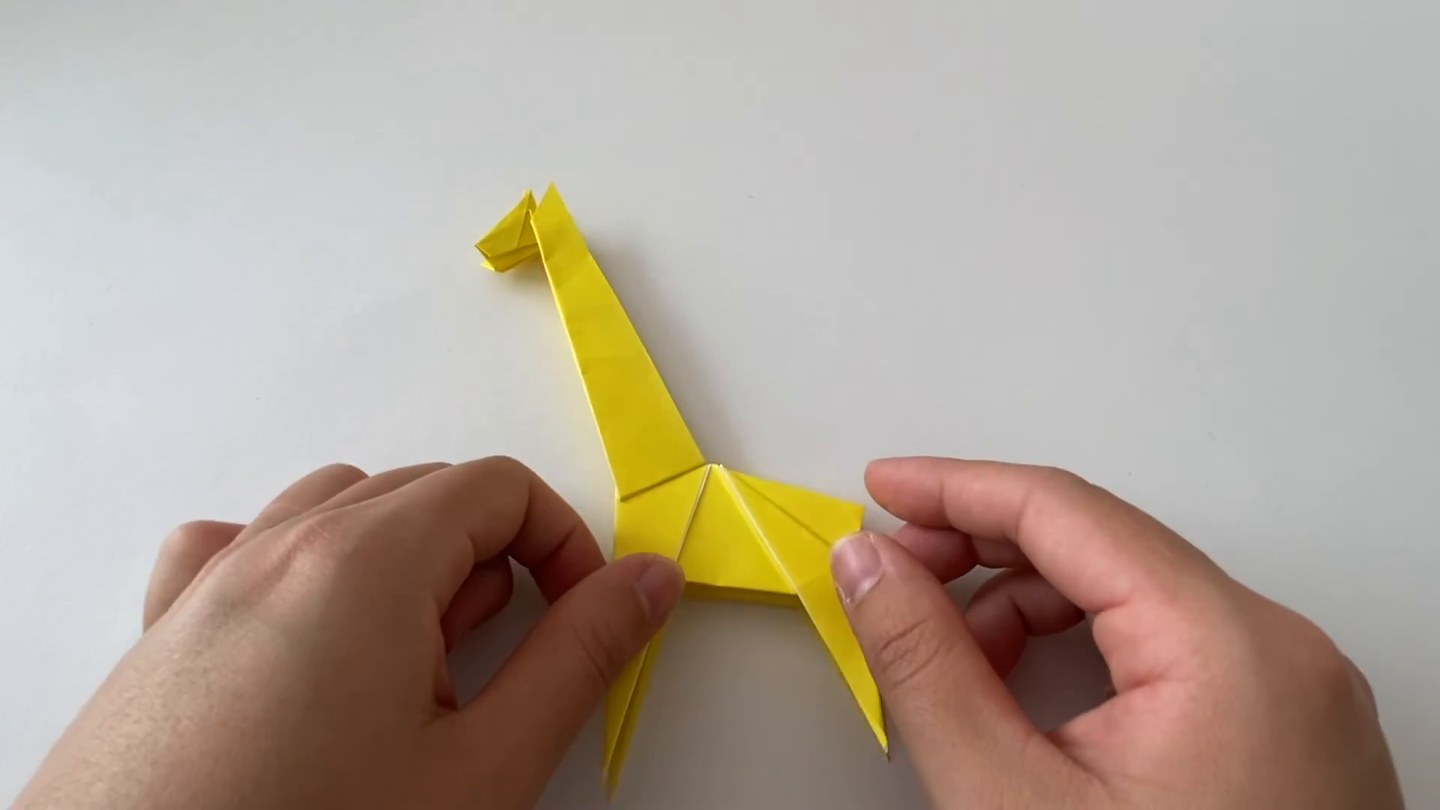 黄色いキリンの折り紙を両手で持っている。首が上に伸び、胴体から脚が出ている