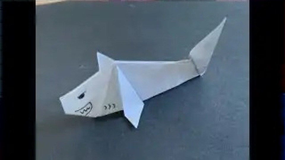 白い折り紙で折ったサメが、ぎざぎざの歯とつり上がった目を描いた顔で、背びれと尾びれを立てて黒い机の上に置かれている