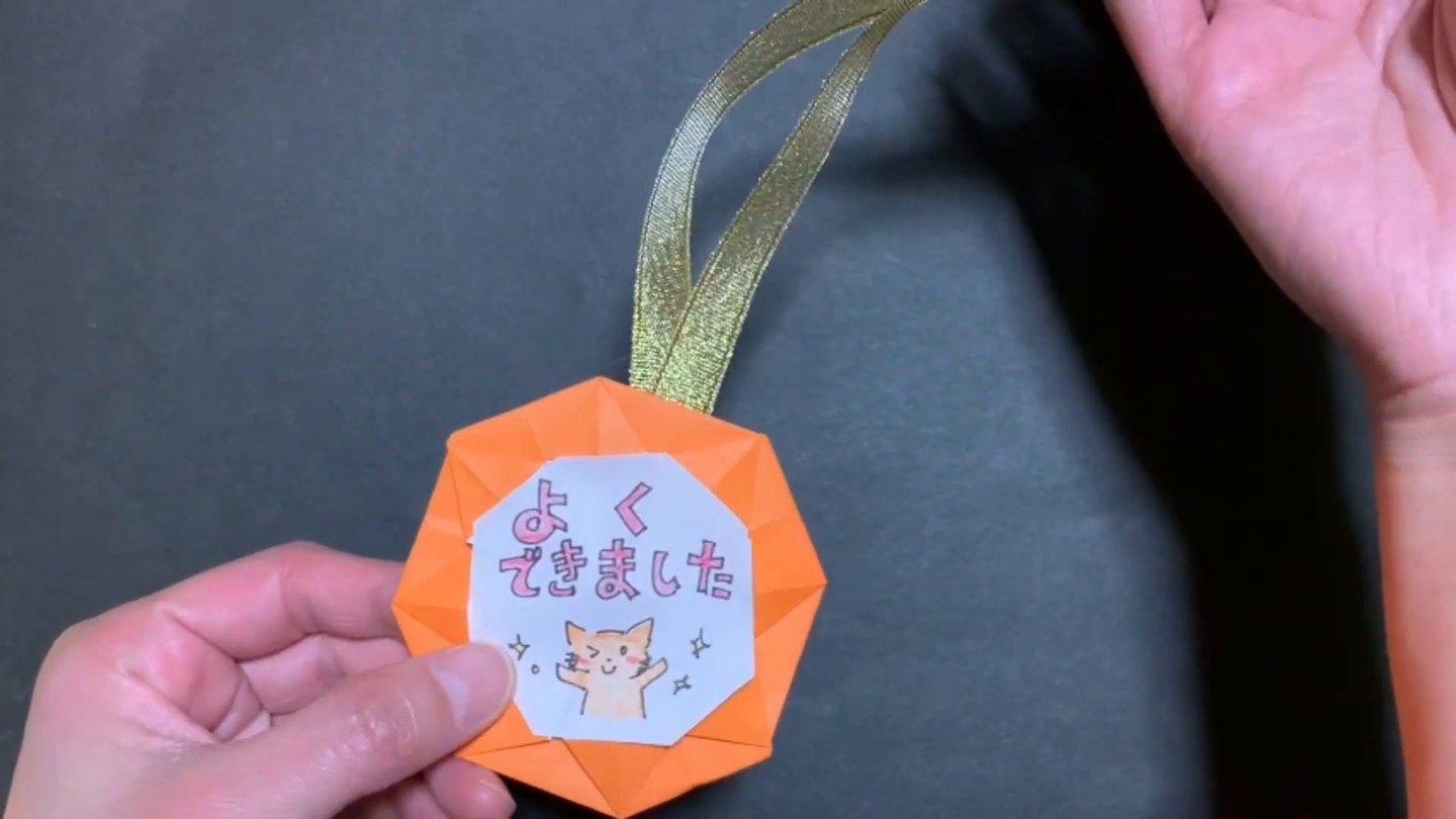 金色のリボンを通したオレンジ色のメダルを手で持ち、中心の白いカードに猫のイラストと文字が見える