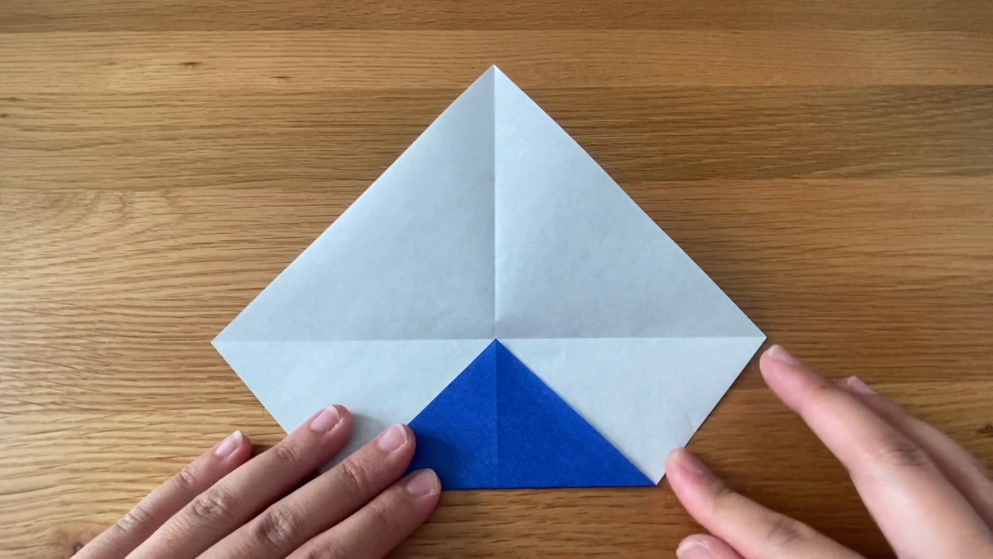 裏の白い面を上にした折り紙の下の角が折り上がり、青い小さな三角が見えているところ