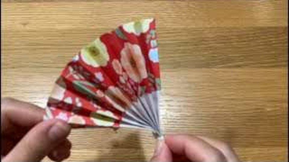 赤い花柄の折り紙を扇形に広げた扇子を、片手で持っているところ